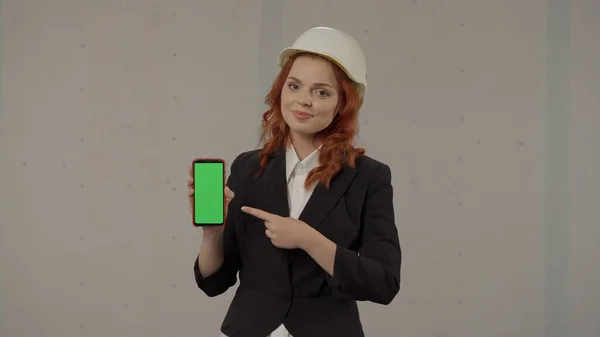 一位女建筑师将智能手机屏幕面向前方 指向绿色屏幕 工作室里一个戴着灰色背景头盔的女商人的画像 广告领域 — 图库照片