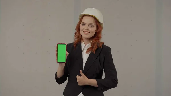 スマートフォンの緑色のスクリーンを見せて親指を上げる建築家 スタジオの灰色の背景にヘルメットとスマートフォンのビジネス女性の肖像画 広告エリア ワークスペース — ストック写真