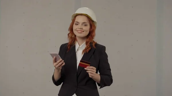 一位女建筑师用智能手机和信用卡在网上商店里买东西 在灰色背景的工作室里 一个戴着保护头盔的女商人的画像 — 图库照片