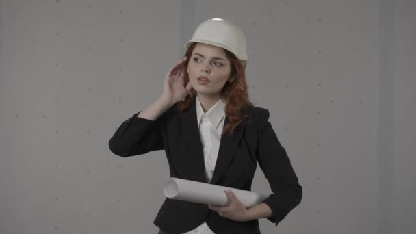 赤毛の女性建築家は耳を傾け 注意深く耳を傾けようとする 灰色の背景にスタジオで彼女の手の青写真でハードハットで女性の肖像画 — ストック動画