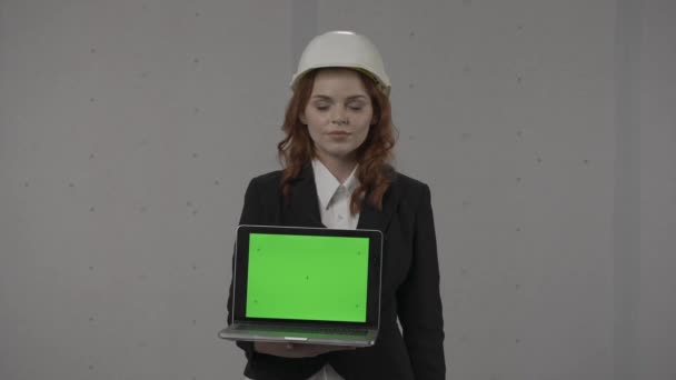 女性アーキテクトが前に向かってラップトップスクリーンを保持し 緑色の画面を指しています スタジオで灰色の背景に彼女の手のハード帽子とラップトップでビジネス女性の肖像画 — ストック動画