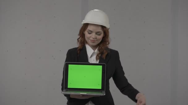 女性建築家が前に向かってラップトップスクリーンを保持し 緑色のスクリーンを指し 親指を上げます ハードハットでビジネス女性の肖像画と彼女の手のラップトップと灰色の背景 — ストック動画