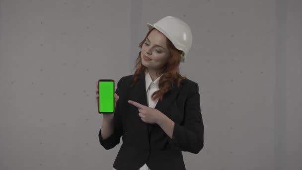 女性の建築家がスマートフォン画面を前面に持ち 緑色の画面を指差しています スタジオの灰色の背景にヘルメットでビジネス女性の肖像画 広告エリア — ストック動画