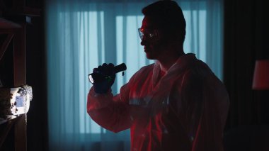 Tulum ve gözlük konusunda genç bir adli tıp uzmanı bir suç mahallini incelerken el feneri kullanıyor. Kırmızı ve mavi polis sirenleriyle aydınlatılmış karanlık bir dairede adli tabip.
