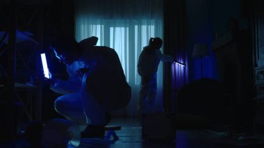 Adli tıp uzmanlarından oluşan bir ekip, polis sirenlerinin mavi ışığıyla aydınlatılan karanlık bir dairede suç mahallinde çalışıyor. Ultraviyole lamba kullanan bir adam ve bir kadın kanıt, parmak izi ve DNA arıyorlar.
