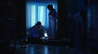 Koruyucu giysiler ve gözlükler içinde adli tıp uzmanlarından oluşan bir ekip kurbanların cesedini bir ceset torbasına koyuyor. Uzmanlar, bir adam ve bir kadın karanlık bir dairede mavi ışıkla aydınlatılmış.