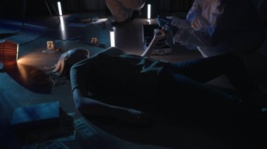 Kadın cesedinin üzerinde eğilen adli tıp uzmanı, kurbanların tırnaklarının altından DNA örneği alıyor, içinde iş malzemeleri olan açık bir evrak çantasının yanında. Bir suç mahallinde koruyucu kıyafetli bir kriminolog.