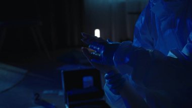 Kadın cesedinin üzerine eğilen adli tıp uzmanı, kurbanların tırnaklarında failin DNA 'sını arıyor. Eldivenli adli tabibin elleri ve karanlık bir dairedeki kurbanın elleri.