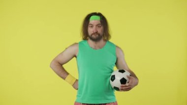 Spor kıyafetli bir adam futbol topu tutuyor, başparmağını kaldırıp kameraya doğrultuyor. Yeşil kolsuz tişörtlü ve bantlı bir erkek portresi. Spor konsepti. Sarı arkaplanda izole edilmiş.