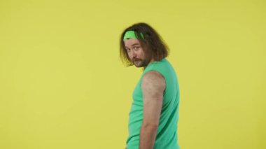 Spor kıyafetli bir adam arkası dönük poz veriyor, yüzünü kameraya çeviriyor ve ekranı işaret ediyor. Yeşil kolsuz tişörtlü ve bantlı bir erkek portresi. Sarı arkaplanda izole edilmiş.