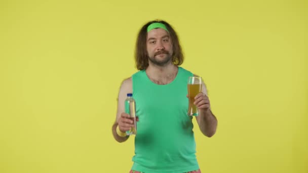 水のボトルとビールのグラスを持っているスポーツウェアの男 チョス 親指を示す 緑のタンクの上およびヘッドバンドの男性の肖像画 黄色の背景に分離されています 健康的なライフスタイルのコンセプト — ストック動画