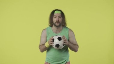 Spor giyimli bir adam futbol topu tutuyor, maç izliyor, takım gol atarken mutlu görünüyor. Yeşil kolsuz tişörtlü ve bantlı bir erkek portresi. Spor konsepti. Sarı arka planda izole edilmiş. HDR BT2020 HLG