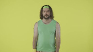 Spor kıyafetli bir adam kameraya negatif yüzüyle bakıyor, başını sallıyor. Yeşil kolsuz tişörtlü ve bantlı bir erkek portresi. Sağlık sigortası konsepti. Sarı arka planda izole edilmiş. HDR BT2020 HLG