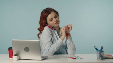 Mutlu ve rahat bir genç kadının orta izole edilmiş fotoğrafı. Telefonla konuşurken tırnaklarını törpüleyip gülümsüyor. Laptop ve çalışma malzemeleri. İş içeriği veya reklam.