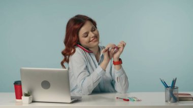 Neşeli ve aktif genç bir kadının, telefonuyla konuşurken ve gülümserken tırnaklarını törpülediği orta ölçekli bir görüntü. Laptop ve çalışma malzemeleri. İş içeriği veya reklam.