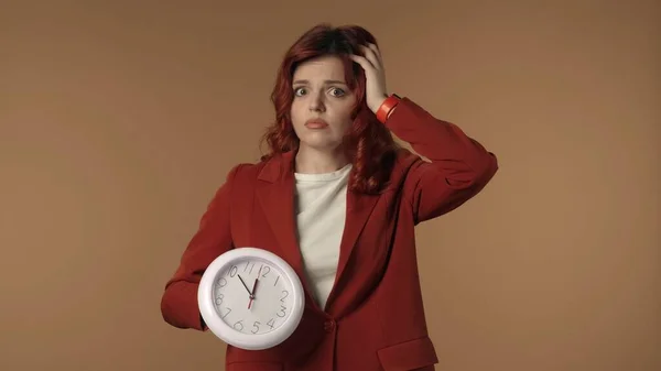 Medium isolated shot of a shocked and worried young woman looking at the clock and holds on to her head. She is confused about the time, being late. Business content or advertisement. Time management.