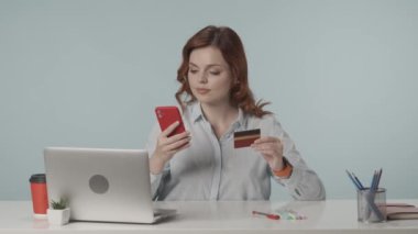 Neşeli, memnun ve rahat genç bir kadının internet üzerinden alışveriş yaparken telefonuna ve kredi kartına baktığı orta ölçekli bir video. İnternet kaynakları. Çevrimiçi hizmetler ve bankacılık reklamı