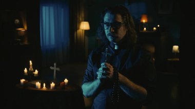 Karanlık odada elinde boncuklar ve elinde haçla gözleri kapalı dua eden bir rahibi yakalarken vuruldu. Arka planda sihirli aletlerle dolu bir masa var. Yaratıcı içerik.