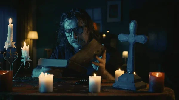 拍到一个牧师坐在桌旁 被蜡烛 十字架和珠子环绕 手里拿着一本书 仔细阅读 这个人在做一个仪式 创意内容 Hallobetween — 图库照片