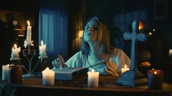 拍到一个年轻女子坐在桌旁 被蜡烛 十字架和珠子环绕 手里拿着一本打开的魔法书 表演着一个仪式 一个仪式 读着魔法 创造性的 介于两者之间的 — 图库照片