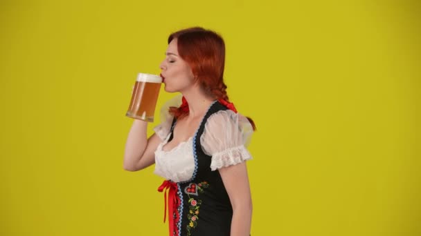 中黄色背景隔离视频 一个年轻的德国女人 女服务员 穿着传统服装 拿着一杯啤酒 喝了一口 咽了口气 舔了舔嘴唇 十月节 啤酒节 — 图库视频影像