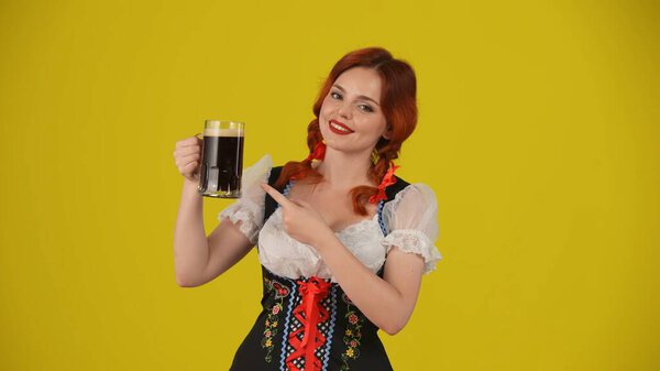Medium yellow background isolated shot of a young German woman, waitress, wearing a traditional costume, holding a glass of dark beer, pointing at it, smiling. Octoberfest, beer festival.