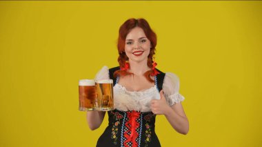 Geleneksel bir kostüm giymiş, elinde bir bardak bira, bira, baş parmak işareti yapıp gülümseyen genç bir Alman kadının orta sarı arka plan görüntüsü. Ekim Festivali, bira festivali.