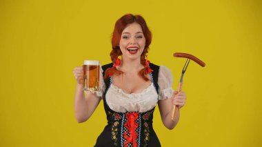 Geleneksel bir kostüm giymiş, elinde sosis ve bir bardak bira tutan, heyecan ve heyecan ifade eden genç bir Alman kadının orta sarı arka plan görüntüsü. Ekim Festivali, bira festivali.