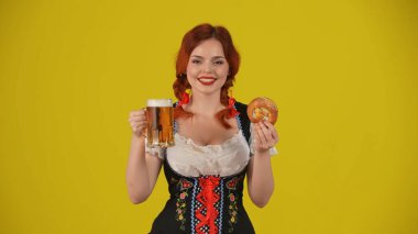 Geleneksel bir kostüm giymiş, elinde çubuk kraker, bir bardak bira ve gülümseyen genç bir Alman kadının orta sarı arka plan görüntüsü. Ekim Festivali, bira festivali, reklam.
