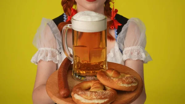 一个无法辨认的年轻德国女人 身穿传统服装的女服务员 手里拿着一个装有椒盐卷饼 乌尔斯和一杯啤酒的盘子 拍了一张黄色背景的离谱照片 10月节 — 图库照片