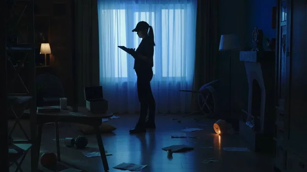 Crime scene creative concept. Police woman with tablet standing in the dark living room. Portrait of female in uniform in the apartment with signs of robbery.