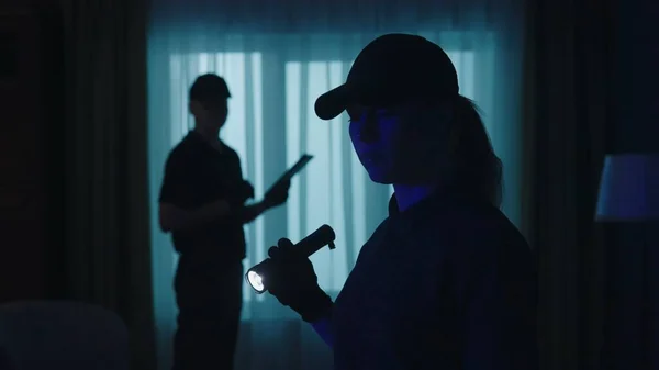 Crime scene creative concept. Focus on the woman with flashlight. Closeup shot of police officers standing in the dark living room, looking around, holding tablet.