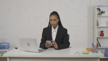 Modern iş kadını yaratıcı konsepti. Ofisteki genç Afro-Amerikan kadın portresi. Masada dizüstü bilgisayarda çalışan, akıllı telefondan mesaj alan odaklanmış bir kadın.