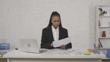 Modern iş kadını yaratıcı konsepti. Ofisteki genç Afro-Amerikan kadın portresi. Kadın evrakları kontrol ediyor ve laptopa bakıyor, hiçbir şey bulamıyor, hayal kırıklığına uğramış bir şekilde iç çekiyor.