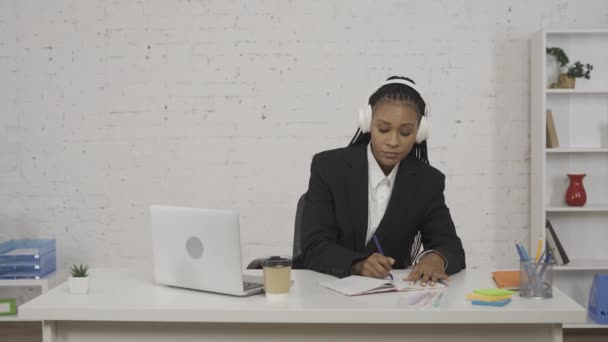 現代のビジネスマンコンセプト オフィスの若いアフリカ系アメリカ人女性の肖像画 ヘッドフォンの女性は音楽を聴き ラップトップで作業し ノートブックで書いたり コーヒーを飲んだり 楽しんだりしています Hdrについて — ストック動画