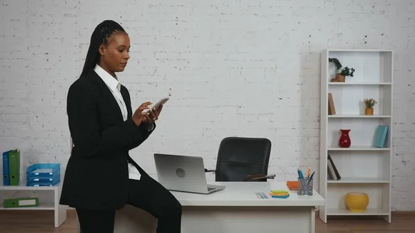 现代女商人的创意概念 办公室里年轻非洲裔美国女性的画像 身穿黑色西服的女性坐在桌子上 手持智能手机 阅读新闻或社交媒体 — 图库照片