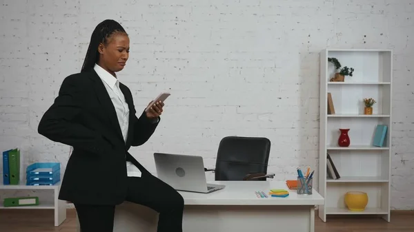 现代女商人的创意概念 办公室里年轻非洲裔美国女性的画像 身穿黑色西服的女人坐在桌子上 拿着智能手机看书 看上去很不高兴 — 图库照片
