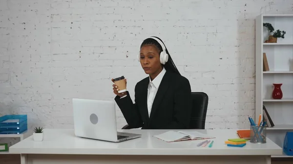 现代女商人的概念 办公室里年轻非洲裔美国女性的画像 坐在办公桌前 头戴耳机 一边听音乐 一边用笔记本电脑 喝咖啡的女人 — 图库照片