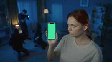Evde suçüstü yakalanan iki hırsızın videosu var. Polis onlara silah doğrultuyor. Ön planda elinde akıllı telefon olan yeşil ekranlı bir kadın var. Muhafızlar