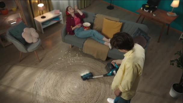 上からのビデオ 床を掃除する若者をキャプチャします ソファーの後ろに横たわる若い女性が携帯電話をスクロールしてトウモロコシを食べて床に落としてしまいました 男は苛立っている — ストック動画