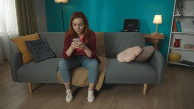 Oturma odasında elinde akıllı bir telefonla kanepede oturan kızıl saçlı genç bir kadının fotoğrafı. Sakin ve rahat görünüyor. Ev, konfor ve konfor. Reklam, yaratıcı içerik.