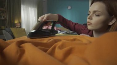 Genç kızıl saçlı bir kadının kapalı mekan videosunu kapat. Kadın giysilerini ütülemek için buhar ütüsü kullanıyor. Odaklanmış görünüyor. Ev aletleri, ev işleri. HDR BT2020 HLG Malzemesi.