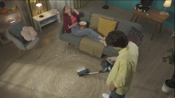 上からのビデオ 床を掃除する若者をキャプチャします ソファーの後ろに横たわる若い女性が携帯電話をスクロールしてトウモロコシを食べて床に落としてしまいました 男はイライラしている Hdrについて — ストック動画