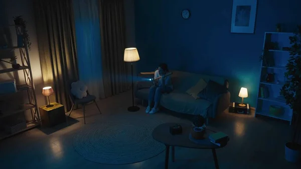 Shot of a young woman sitting on a couch in a dark room looking around for intruders. She holds a baseball bat and a flashlight. Safety, guard or security agency, advertisement.