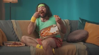 Şişko, orta yaşlı bir adam kanepeye bacağı üzerinde oturur. Adam büyük bir zevkle iki pizza yiyor. Odada bazı spor aletleri var. Zararlı yiyecekler. Vücut pozitifi.