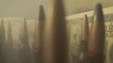 Dolar banknotlarının arka planındaki fişekler dumanlı bulutlara yakın. Mercek altında silah satışı, silahlara yardım, orduda çalışmak, mühimmat ticareti.