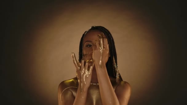 黄金の唇 黄金の肌 黄金の鎖を持つ女性は 円形のライトでブラウンの背景にスタジオで彼女の髪に アフリカ系アメリカ人女性の腕の上に液体の金が流れる — ストック動画