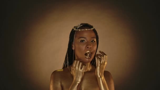 非洲裔美国妇女在克利奥帕特拉风格的棕色背景与圆形光 一个头上戴着金色皮肤和珠宝的女人嘴里衔着一颗含金巧克力糖 — 图库视频影像