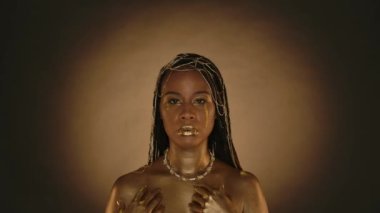 Yüzünde altın gözyaşı damlaları olan Afrikalı Amerikalı bir kadının portresi. Altın metalik makyajlı, kahverengi arka planda mücevherleri ve dairesel ışığı olan örnek bir kadın. Moda.