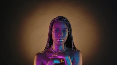 Yüzünde pembe neon ışıkta altın gözyaşı damlaları olan bir kadının portresi. Altın metalik makyajlı, kahverengi arka planda mücevherleri olan, Afro-Amerikalı bir kadın.